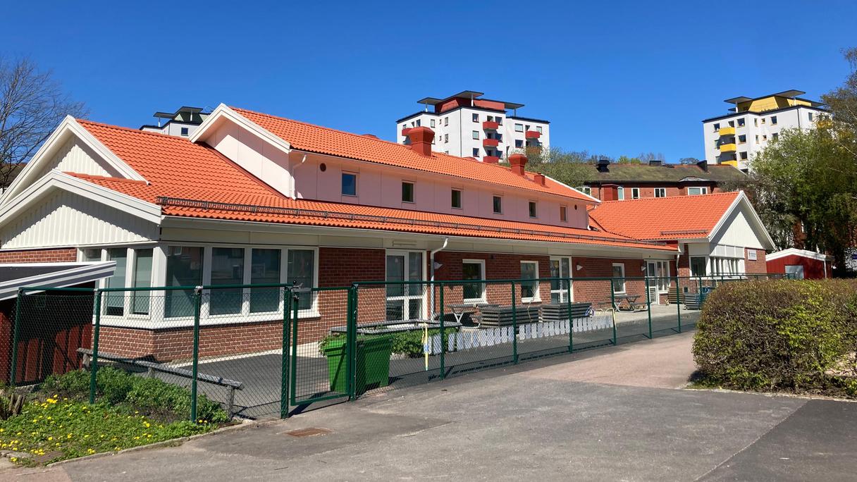 Bilden föreställer ett tegelhus med ingång till förskolans avdelningar. Ett staket ramar in byggnaden.