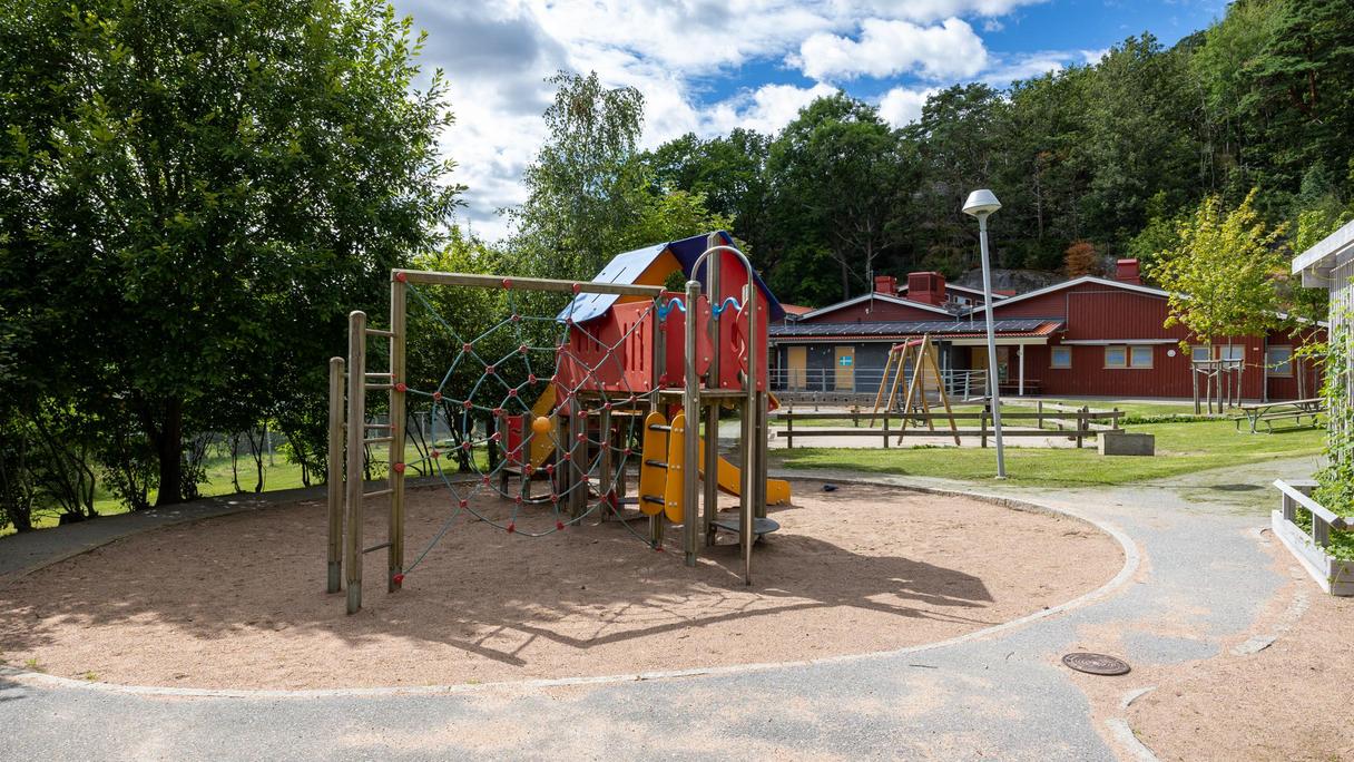 Bilden föreställer en röd byggnad och en förskolegård. På gården finns klätterställningar och träd.