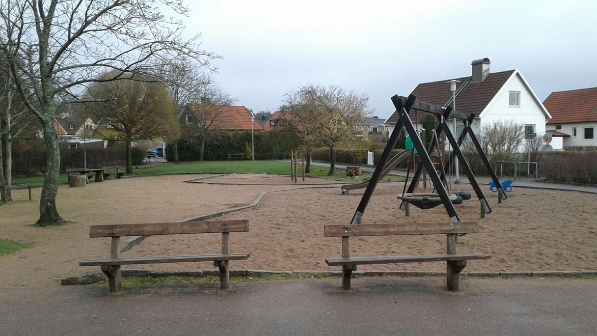 Lekplats i bostadsområde med träd, gräsmattor, bänkar och sandlådor med lekredskap