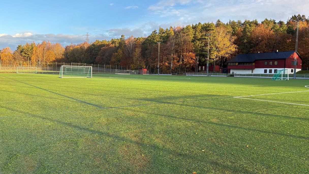 Grön bollplan i solsken. I bakgrunden syns en klubbstuga