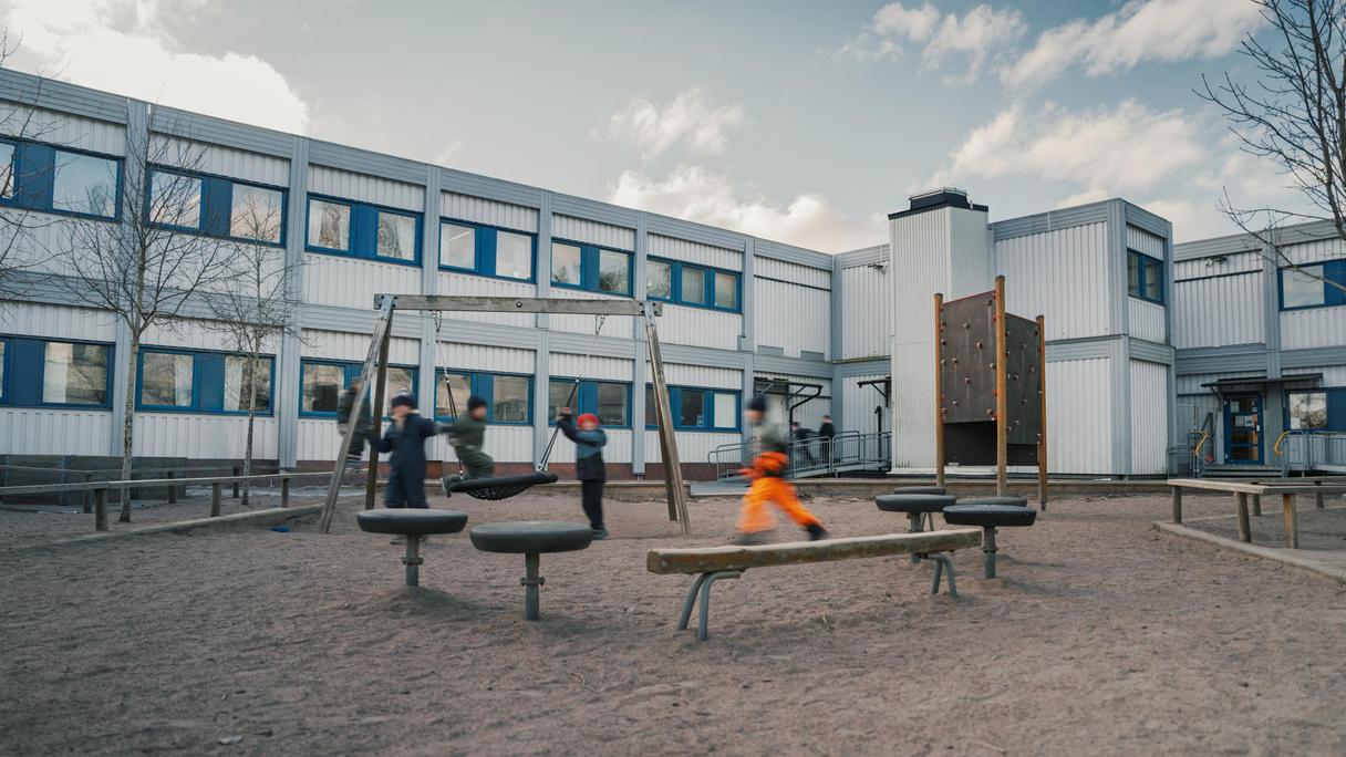 En grupp elever som leker på en skolgård med en sandlåda och en vit skolbyggnad i bakgrunden.