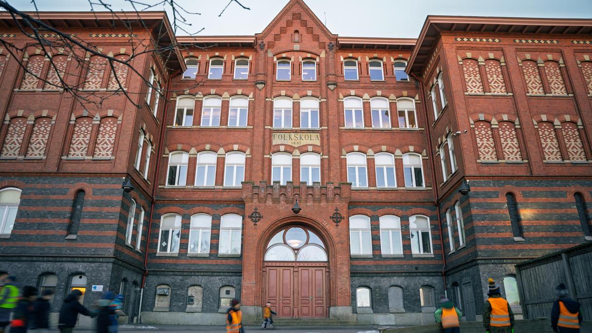 En grupp elever som står på skolgården framför en en stor skolbyggnad i rött tegel.