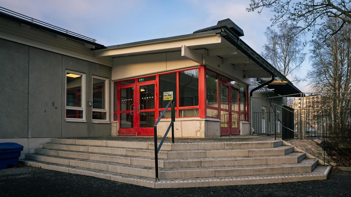 En grå skolbyggnad med röd entré med trappor som leder upp till den.
