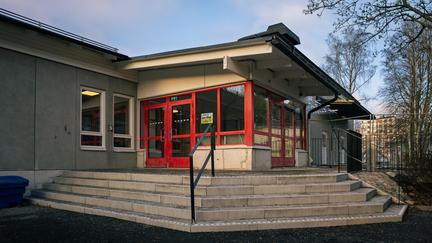 En grå skolbyggnad med röd entré med trappor som leder upp till den.