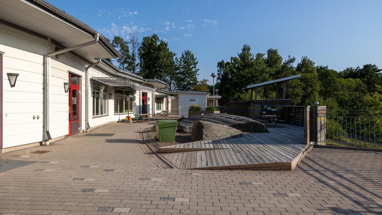 Bilden föreställer en vit byggnad med en röd dörr. Utanför byggnaden finns en uteplats.