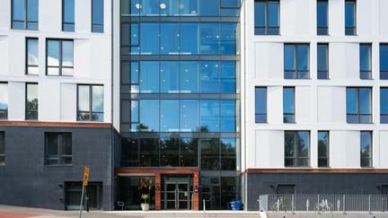 Ett modernt flerbostadshus med en stor glasfasad i mitten och vita fasader med många fönster på sidorna. Entrén ligger vid gatan framför huset.