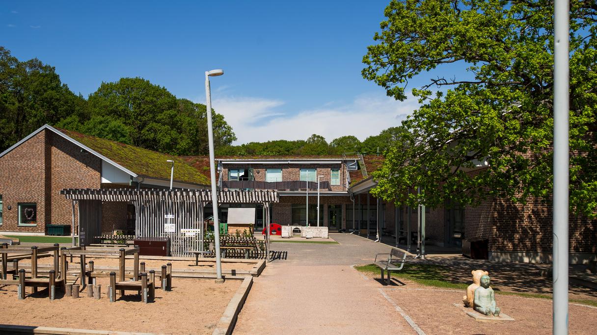 Förskolan i brunt tegel och förskolans gård med sandlådor, pergola och klätterställningar.