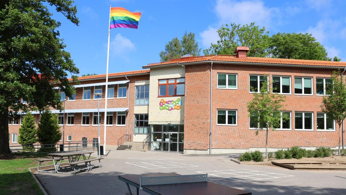 Utbynässkolans huvudentré. Solen skiner, himlen är blå och en pride-flagga är hissad på flaggstången. Bladen på träden är gröna och det står ett pingisbord på skolgården.
