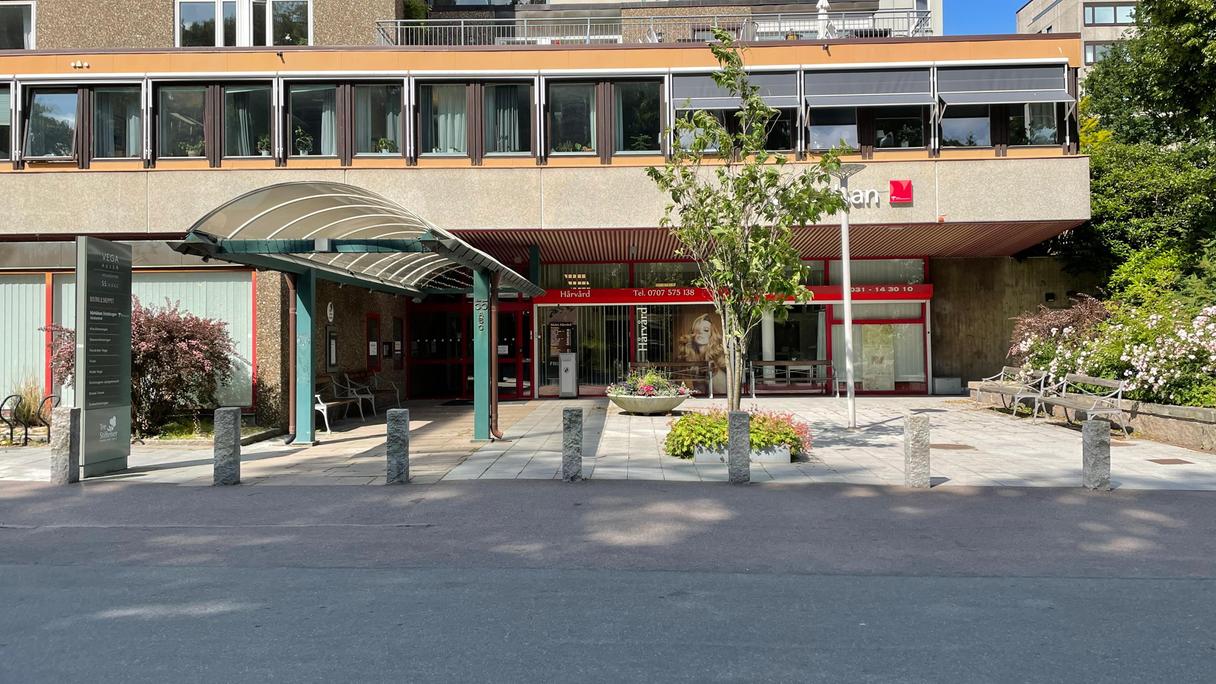 Entrén till Vegahusen ligger i en grå byggnad på marknnivå. Marken är platt och framför ingången finns två bänkar.