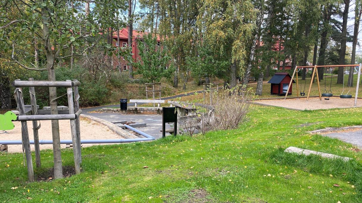 Lekplatsen ligger i en skogsdunge med sluttande gräsmattor, bänkar och sandlådor med lekredskap
