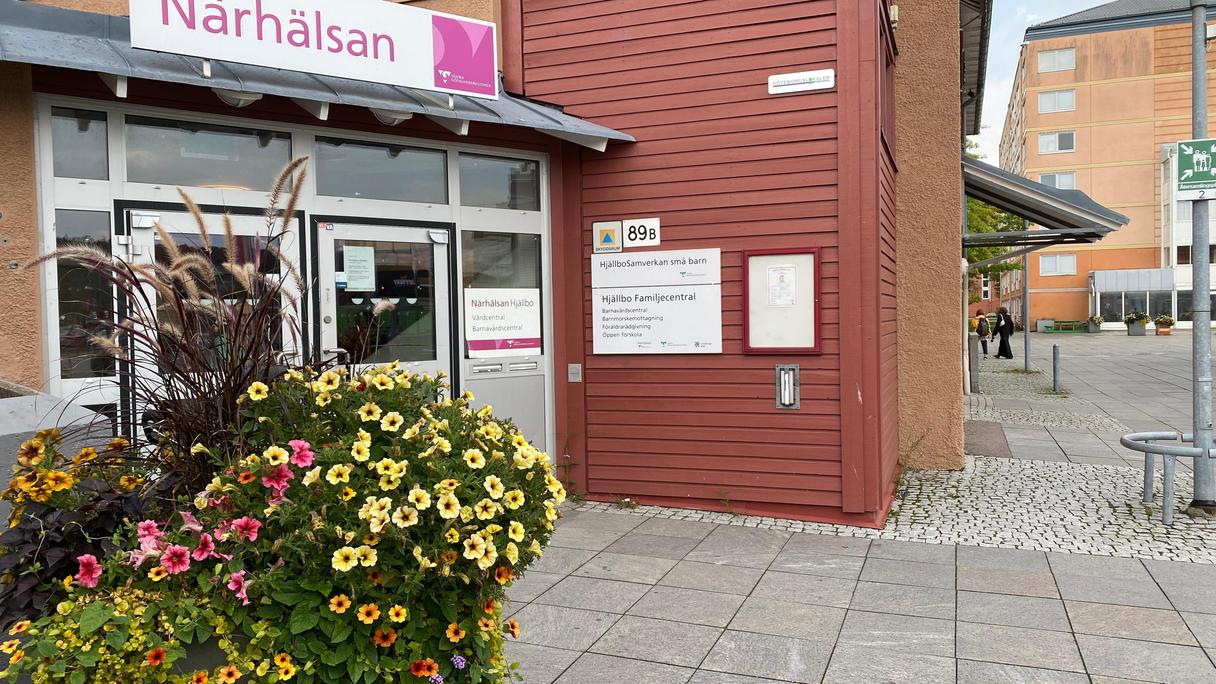 En röd dörr i ett två våningshus med skylt Närhälsan. Framför står ett blomsterarrangemang. st