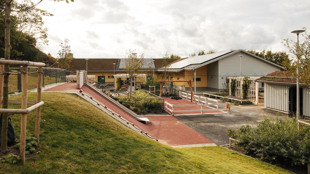 Bilden föreställer en gul och grå byggnad. Framför byggnaden finns en lekplats med en gunga, en ruschkana och utomhusbänkar. 