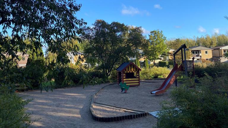 Lekplatsen har sand som underlag med grönska runt om och bänkar att sitta på