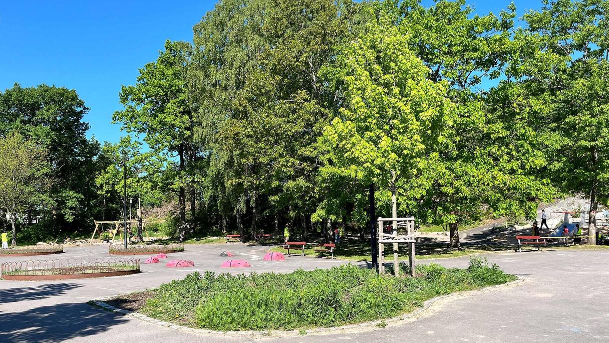 En lekplats med lummiga träd, gungor, hårdgjorda ytor och rabatter