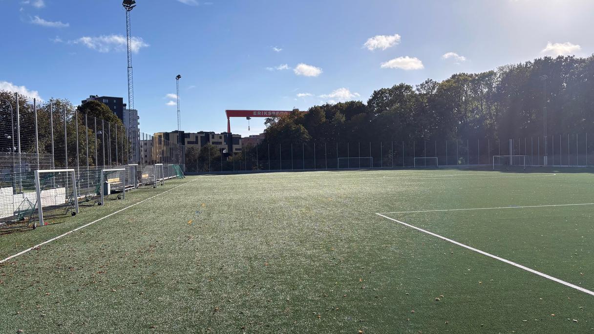En grön fotbollsplan under blå himmel. I bakgrunden syns Eriksbergskranen,