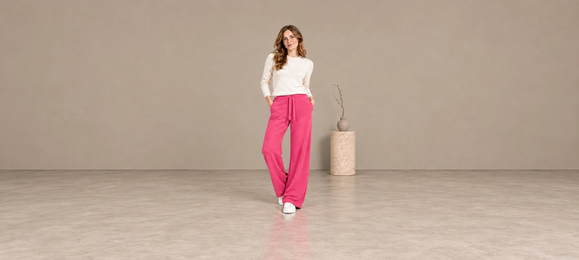 Woman in an ivory sweater and pink wide-leg knit trousers beside a stone pedestal and vase