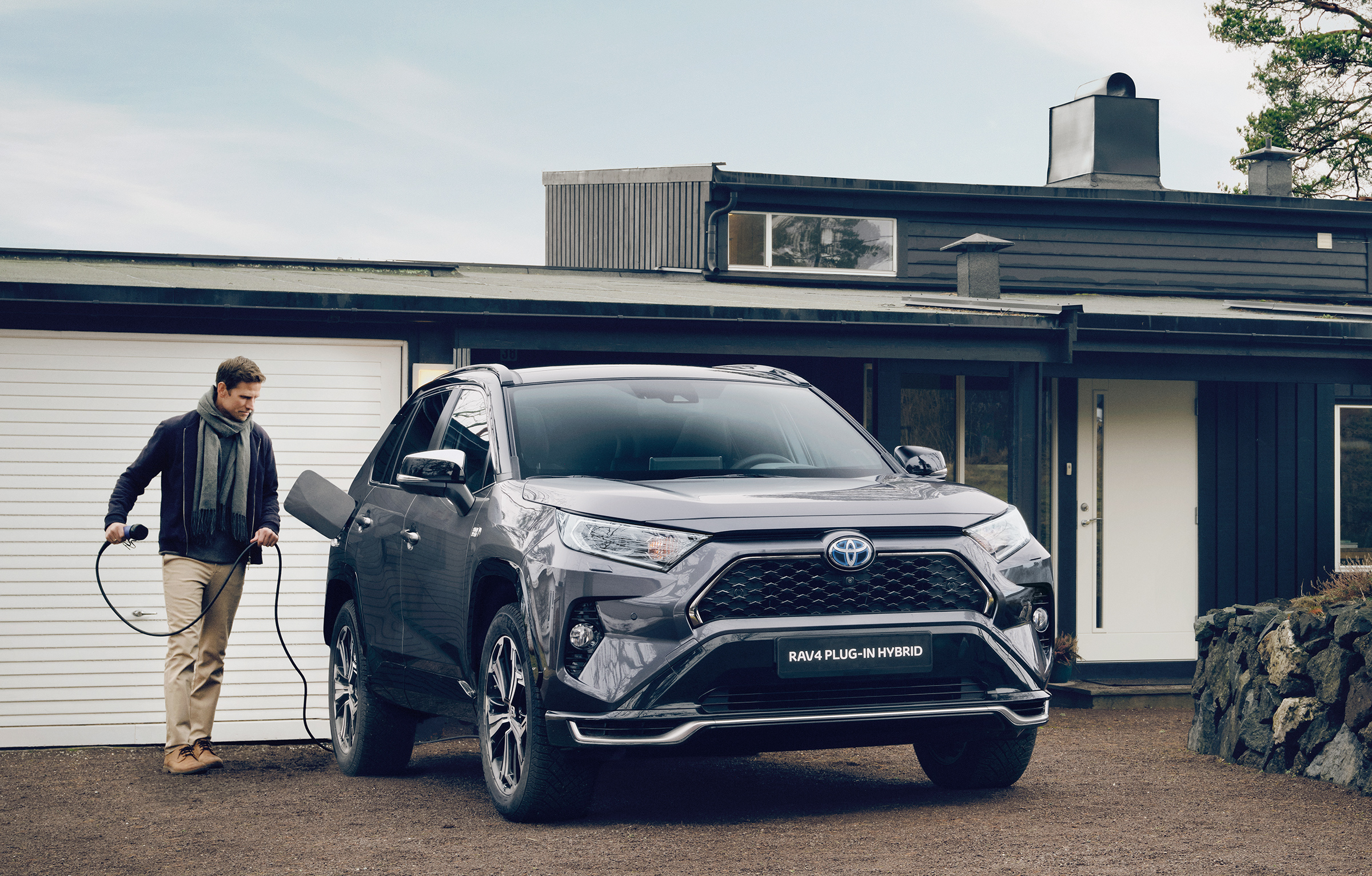 En Toyota RAV4 Plug-in Hybrid står parker i oppkjørelsen til en enebolig. En voksen mann plugger laderen i bilen .